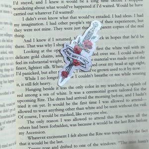 May include: A sticker featuring a dagger with red floral accents and a banner that reads "WE WILL RISE." The sticker is placed on a book page with printed text.