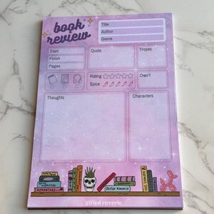 May include: A pastel pink book review notepad with a bookshelf illustration at the bottom. The notepad has sections for title, author, genre, and more. The text "book review" is at the top in a whimsical font.