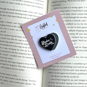May include: A black heart-shaped enamel pin with the words "Shadow Daddy" in white script. The pin is on a pink card with the Gifted Reality logo and website address. The background is an open book.