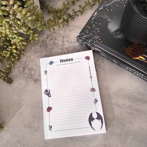 May include: A white notepad with lined pages and decorative floral and bat wing illustrations. The notepad has the word "Notes" printed at the top. A book and potted plants are in the background.