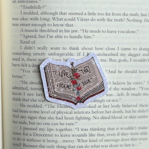 May include: A sticker featuring an open book design with the text "I LOVE THAT FUCKING BOOK." The book illustration is in shades of brown and red, with a sword and red flowers. The sticker is on a white background.