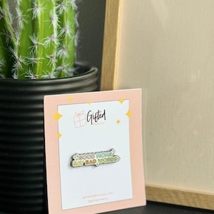 May include: A pin with the text "GOOD MOMS SAY BAD WORDS" on a pink card with a gold frame and a cactus in the background. The pin is green, white, and black. The card has the Gifted Reality logo.