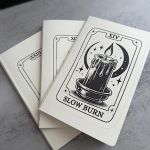 May include: Three off-white rectangular notebooks with black ink illustrations. The top notebook features a tarot-style design of a lit candle in a holder, a crescent moon, stars, the Roman numeral XIV, and the text SLOW BURN at the bottom.