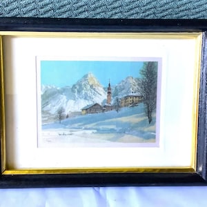 May include: A framed print depicting a winter scene with a church steeple and snow-capped mountains. The print is in a black frame with a gold inner border.