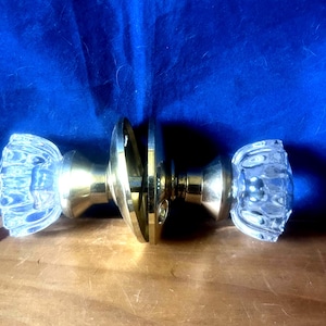 May include: A vintage door knob set with clear, faceted glass knobs and brass hardware. The knobs are symmetrical, with a decorative, ribbed design. The set includes a central plate and connecting hardware.