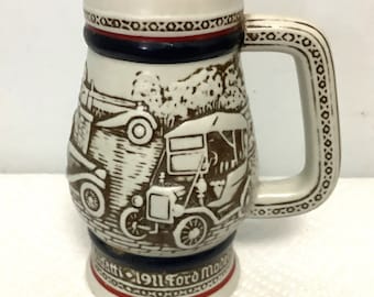 May include: A white ceramic beer stein with a brown and white image of a 1911 Ford Model T car. The stein has a blue and red band around the top and bottom. The text "1911 Ford Model T" is printed on the bottom of the stein.