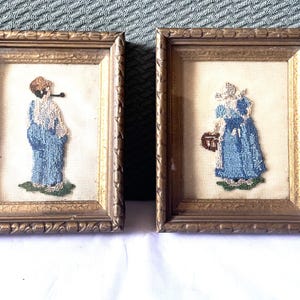 May include: Two framed needlepoint embroidery pieces with gold frames. The embroidery depicts two figures in blue dresses.
