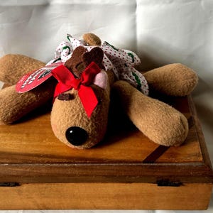 May include: A tan plush reindeer toy with a black nose, wearing a white dress with a red and green pattern and a red ribbon bow on its head. A red tag is attached to the reindeer, which is positioned on a rectangular wooden box.