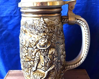 Puede incluir: Un jarro de cerveza vintage con una escena en relieve detallada de un minero de oro, una cabaña y una cascada. La jarra de cerámica tiene una tapa y un asa metálicas con bisagras, con una base y un asa de color crema.
