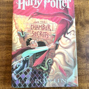 May include: A hardcover book titled "Harry Potter and the Chamber of Secrets" with a vibrant cover illustration. The cover features a red dragon-like creature, a young boy with glasses, and the author's name, J.K. Rowling.
