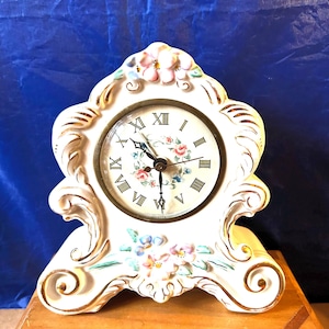 May include: A white ceramic mantel clock with floral and gold accents. The clock face has Roman numerals and a floral design. The clock is decorated with pink, blue, and white flowers.
