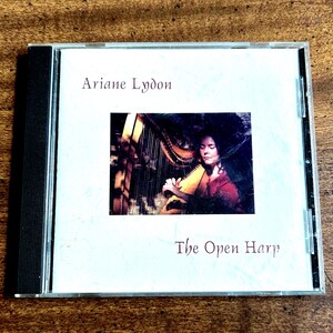 May include: A CD case with the album "The Open Harp" by Ariane Lydon. The cover art depicts a woman playing a harp, with a warm colour palette. The text "Ariane Lydon" and "The Open Harp" are displayed in a serif font.