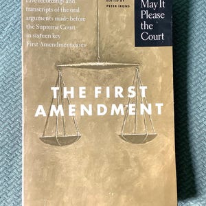 Puede incluir: Una portada de libro con fondo marrón y una ilustración de una balanza. El título es "The First Amendment" y el subtítulo es "May It Please the Court".
