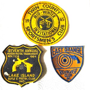 May include: Three vintage sports club patches. The top patch is yellow with black text and a crossed pistol design. The bottom left patch is black with yellow text and pistol graphics. The bottom right patch is orange with blue text and a pistol graphic.