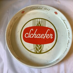May include: Vintage Schaefer beer serving tray. The round metal tray features a white background with a red and green central design. The text "Schaefer" is in white script, and the rim reads "America's Oldest Lager Beer" and "Established 1842."