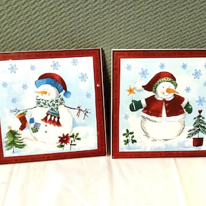 Puede incluir: Dos azulejos de cerámica con temática navideña que representan a muñecos de nieve. El azulejo de la izquierda tiene un muñeco de nieve con un sombrero y una bufanda azules, sosteniendo una media roja. El azulejo de la derecha tiene un muñeco de nieve con un sombrero y un abrigo rojos, sosteniendo una estrella.