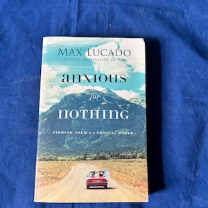May include: A paperback book titled Anxious for Nothing by Max Lucado. The cover features a landscape with a mountain range under a blue sky, a dirt road, and a small red convertible car driving away. The text includes the author's name and subtitle.