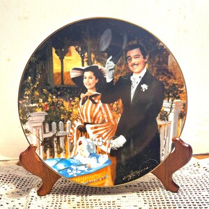 May include: Decorative plate featuring a couple in period attire, with a baby in a basket. The plate has a gold rim and is displayed on a wooden stand. The artwork depicts a scene with a white picket fence and lush greenery.