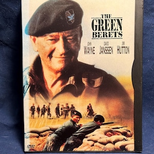 Puede incluir: Caja de DVD de la película "Boinas Verdes" con John Wayne. La portada muestra a Wayne con un boina verde, con el título de la película y los nombres del elenco. El fondo representa soldados en combate.