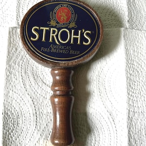 Puede incluir: Un tirador de cerveza de madera con un emblema circular azul, un logotipo de león dorado y el texto STROH'S AMERICA'S FIRE-BREWED BEER. El mango tiene un acabado de madera oscura pulida con una virola de metal dorado en la base y un remate superior dorado.