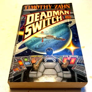 Peut inclure: Un livre de poche intitulé "Deadman Switch" de Timothy Zahn. La couverture présente une scène spatiale avec un vaisseau spatial, une planète avec des anneaux et une personne dans un cockpit. Le livre est un roman de science-fiction.