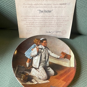 Puede incluir: Un plato de cerámica con una pintura de Norman Rockwell titulada "The Painter". La pintura representa a un hombre arrodillado en un suelo de madera, pintando una pared mientras una niña lo observa. El plato forma parte de la colección Rockwell Heritage.
