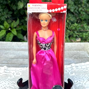 May include: A collectible 11.5-inch fashion doll in a red box. The doll has blonde hair, blue eyes, and wears a bright pink floor-length gown with a silver and black patterned bodice and a small silver necklace. The box features a warning label.