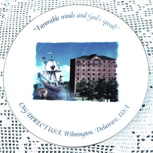 May include: A white decorative plate with a gold rim featuring a nautical scene with a ship and a brick building. The plate has the text "Favorable winds and God's speed!" and "ING DIRECT USA, Wilmington, Delaware, USA".