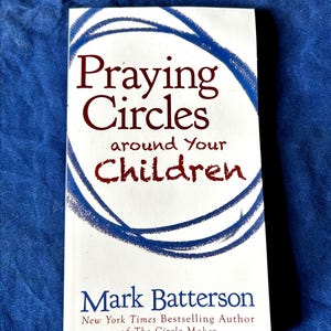 以下が含まれることがあります： Praying Circles around Your Childrenというタイトルの白いブックカバー。著者はニューヨーク・タイムズのベストセラー作家Mark Battersonで、The Circle Makerの著者でもあります。カバーには、濃い赤色のテキストを囲む手描きの青い円形のグラフィックが描かれています。