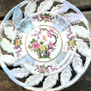 May include: A decorative porcelain plate with a floral design. The plate features a central image of pink and yellow flowers, a butterfly, and branches. The outer rim has a leaf-like design with gold accents.
