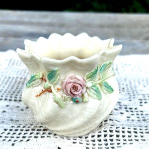 May include: A small, cream-colored ceramic vessel with a scalloped rim and a textured, shell-like body. The exterior is decorated with a three-dimensional relief of a single pink rose surrounded by light green leaves and small blue accents.