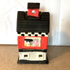 May include: A ceramic farmhouse-style candle holder with a red roof, black and white checkered trim, and a window with a cow and pig peeking out.