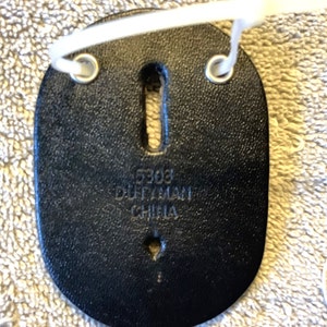 May include: Black leather badge holder with two metal grommets and a keyhole-shaped opening. The text "5303 DUTYMAN CHINA" is embossed on the surface. A white plastic tie is attached to the grommets.
