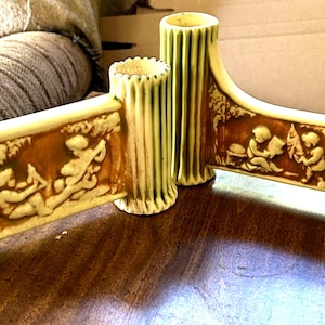 Puede incluir: Una pieza decorativa de cerámica color crema con un diseño en relieve marrón. La pieza tiene cuatro soportes cilíndricos y una sección central con un relieve de figuras. La pieza está sobre una superficie de madera.