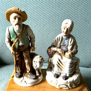 May include: Two ceramic figurines depicting an elderly shepherd and his wife. The shepherd wears a hat, green vest, and carries a staff. His wife is seated, holding a basket. The figurines are painted in muted tones.