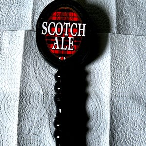 May include: A black Samuel Adams Scotch Ale beer tap handle featuring a circular top with a red and black plaid pattern, white text, and a gold-colored finial at the top and a gold-colored base. The handle has a ridged, cylindrical design.
