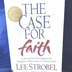 May include: A book titled "THE CASE FOR faith" by Lee Strobel. The cover is white with blue and red text. A gold medallion award seal is in the upper left corner. The book is on a black stand.