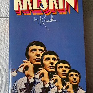Pode incluir: Um livro de bolso vintage intitulado The Amazing World of Kreskin por Kreskin. A capa azul apresenta uma sequência fotográfica repetida de um homem segurando óculos, com letras amarelas e laranjas e o texto The spellbinding mentalist tells all.