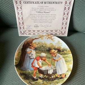 May include: A vintage porcelain plate featuring a painting of three children in a park setting. The children are dressed in colorful clothing and are playing with a doll carriage. The plate is trimmed in gold and has a signature at the bottom.