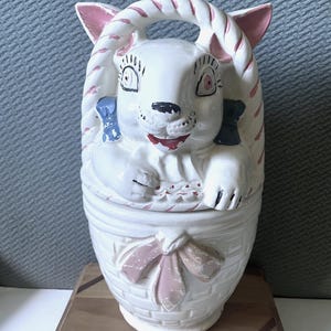 Pode incluir: Jarro de biscoitos de cerâmica branca em forma de coelho numa cesta. O coelho tem orelhas cor-de-rosa, uma pega listrada rosa e branca e laços azuis. A cesta tem um laço cor-de-rosa e um padrão tecido branco e rosa.