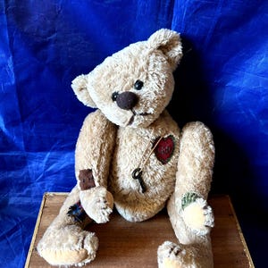 Peut inclure: Un ours en peluche beige avec un nez marron et des yeux noirs. L'ours a un patch en forme de cœur rouge avec une clé et un patch marron sur le bras. Il est assis sur une surface en bois sur fond bleu.