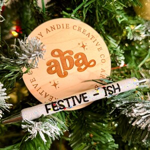 May include: A wooden Christmas ornament with the text "aba" and "By Andie Creative Co." A glittery pen with the text "FESTIVE - ISH" rests on the ornament. The background is a decorated Christmas tree.