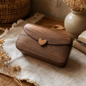 May include: A rectangular dark brown wooden box with a smooth, polished finish and rounded corners. The lid features a curved edge and is secured by a small, centered wooden heart-shaped clasp. The wood grain is visible across the entire surface.