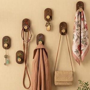 May include: Wooden wall hooks with floral patterns and brass fixtures. A scarf, purse, coat, leash, and keys hang from the hooks. The hooks are mounted on a light beige wall. The items are displayed in a decorative arrangement.