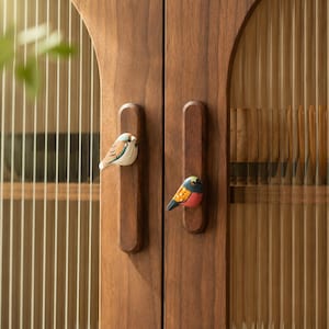 May include: Two wooden cabinet handles mounted on a dark wood cabinet door with fluted glass panels. Each handle features a small, hand-painted bird figurine attached to the front. One bird is white and brown, while the other is black, orange, and red.