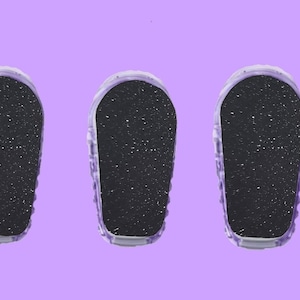 Könnte beinhalten: Drei Fußfeilen mit einer dunklen, strukturierten Oberfläche und einem transparenten, geriffelten Kunststoffrahmen. Die Fußfeilen sind auf einem einfarbigen lila Hintergrund angeordnet. Diese Feilen sind für die Fußpflege bestimmt.