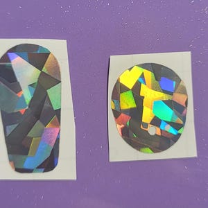 Peut inclure: Deux autocollants irisés sur une surface violette. L'un est de forme ovale, l'autre est un cercle. Les deux autocollants ont un motif géométrique avec un arc-en-ciel de couleurs, dont le vert, le jaune et le bleu.