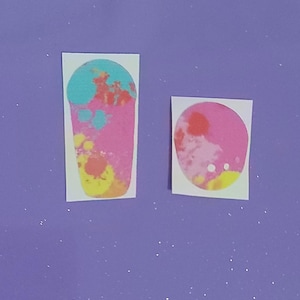 Puede incluir: Dos pegatinas con diseños abstractos coloridos. Una es de forma rectangular alta con salpicaduras de pintura rosa, amarilla y azul turquesa. La otra es una pegatina ovalada más pequeña con colores similares. Ambas están sobre un fondo morado.