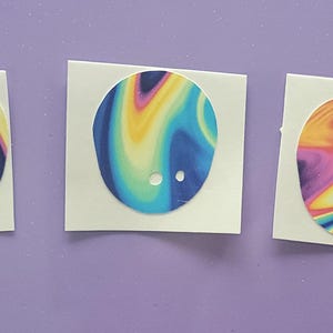 Puede incluir: Tres pegatinas circulares coloridas con un diseño abstracto y arremolinado en tonos de azul, morado, amarillo y rosa. Cada pegatina está sobre un respaldo cuadrado blanco y tiene dos pequeños agujeros. Las pegatinas se muestran sobre una superficie púrpura.