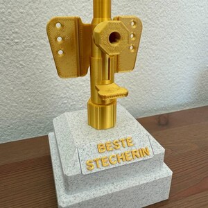 May include: A gold-colored trophy with a white base. The base has the words "BESTE STECHERIN" inscribed on it.